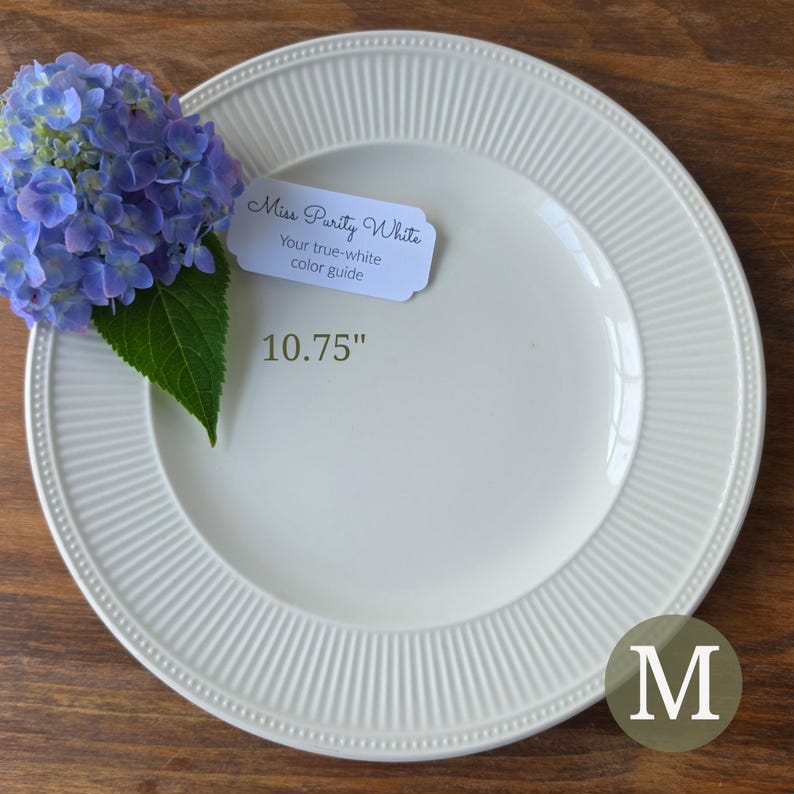 May include: A round white ceramic dinner plate with a ribbed rim and beaded edge, measuring 10.75 inches in diameter. A small white tag reads Miss Purity White Your true-white color guide. A cluster of blue hydrangea flowers rests on the plate.
