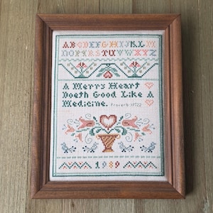 May include: A framed cross-stitch sampler featuring a multi-colored alphabet, floral motifs, and the text A Merry Heart Doeth Good Like A Medicine. Proverb 17:22. The design includes a central heart, a vase, birds, and the year 1989 in a wooden frame.
