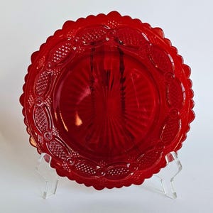 May include: A circular red glass plate featuring a scalloped edge and an intricate embossed pattern. The design includes a central sunburst motif surrounded by a border of textured diamond shapes and raised circular accents on the rim.