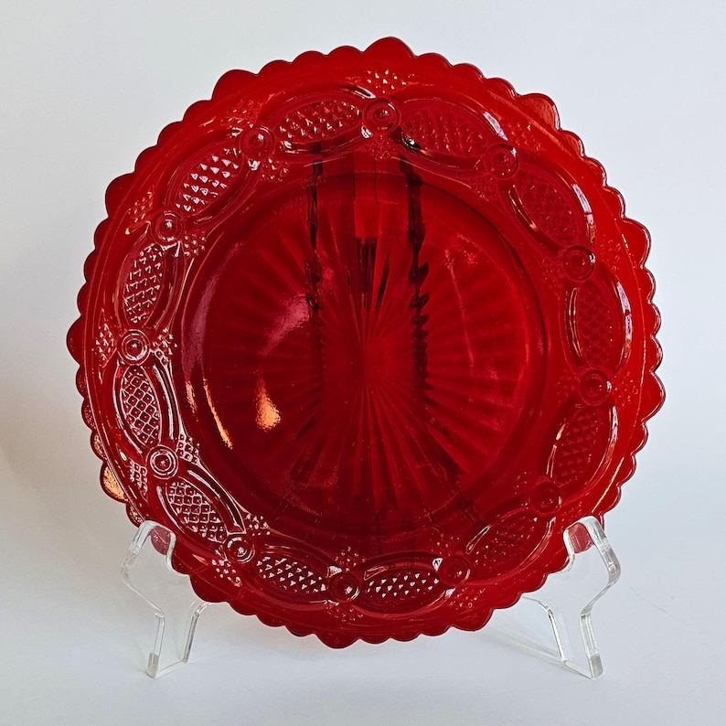 May include: A circular red glass plate featuring a scalloped edge and an intricate embossed pattern. The design includes a central sunburst motif surrounded by a border of textured diamond shapes and raised circular accents on the rim.