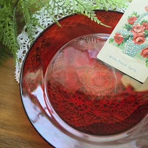 May include: A red glass plate with a decorative pattern, topped with a smaller clear glass dish. A vintage postcard with roses and the text "With Fond Love" rests on the plate. The scene is accented with greenery and lace.