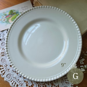 May include: A round, off-white ceramic dinner plate with a smooth, glossy center and a decorative, raised beaded edge detail. The plate is clearly marked with the text 9" indicating its diameter, and the letter G is visible in the corner.