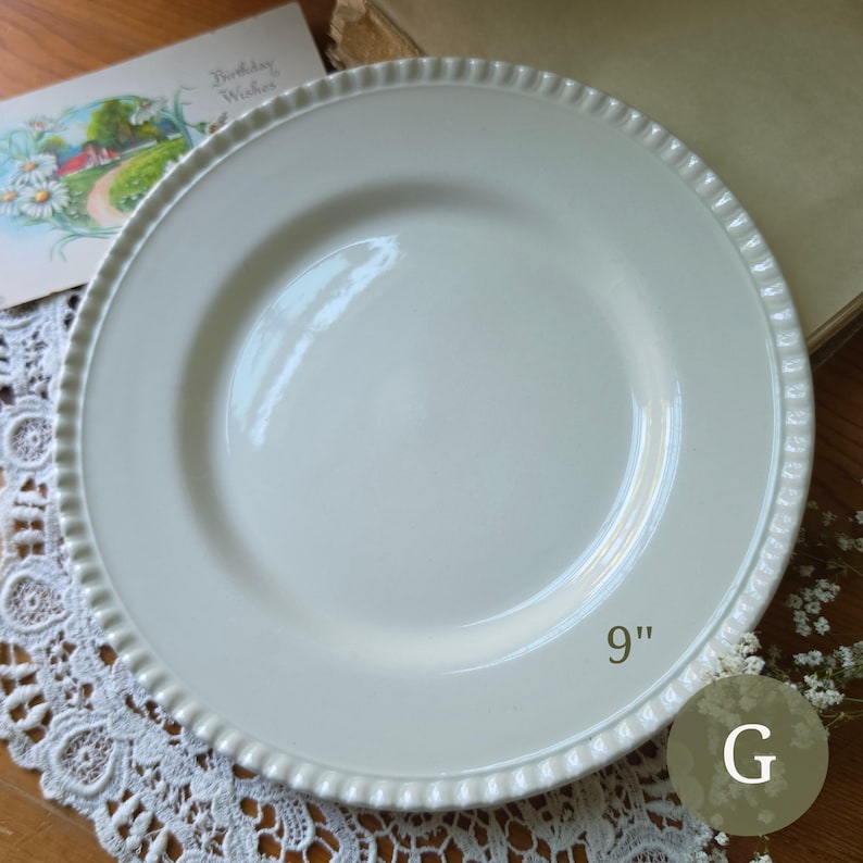 May include: A round, off-white ceramic dinner plate with a smooth, glossy center and a decorative, raised beaded edge detail. The plate is clearly marked with the text 9" indicating its diameter, and the letter G is visible in the corner.