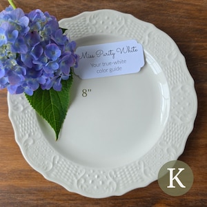May include: An 8-inch off-white ceramic plate with a scalloped, embossed rim featuring a basket-weave and scroll pattern. A label reads Miss Purity White Your true-white color guide. A cluster of purple hydrangea flowers rests on the plate.