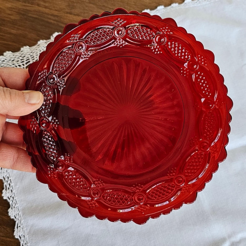 May include: A round, ruby red glass plate featuring a scalloped edge and a decorative border with embossed diamond and oval patterns. The center of the plate displays a sunburst design with radiating lines extending outward from the middle.