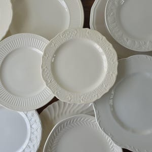 May include: A collection of white ceramic plates featuring various embossed patterns, including scalloped edges, basket weave textures, ribbed rims, and leaf motifs. The assortment displays multiple circular designs with smooth centers and decorative borders.