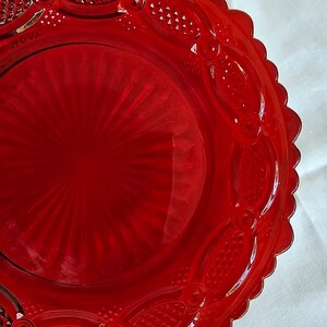 May include: A vibrant red glass plate featuring a central sunburst pattern surrounded by an outer rim with embossed oval shapes and a scalloped edge. The word "ROVA" is clearly visible in raised lettering on the surface of the glass plate.