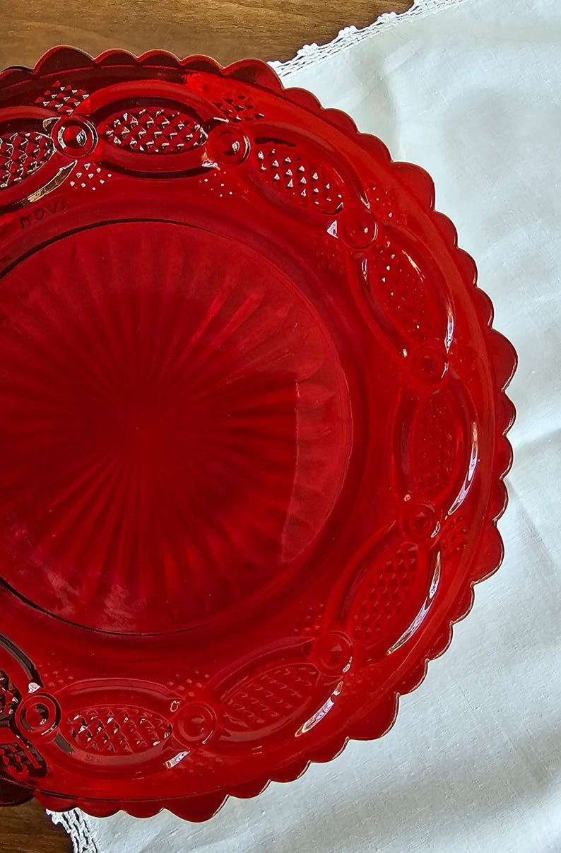 May include: A vibrant red glass plate featuring a central sunburst pattern surrounded by an outer rim with embossed oval shapes and a scalloped edge. The word "ROVA" is clearly visible in raised lettering on the surface of the glass plate.