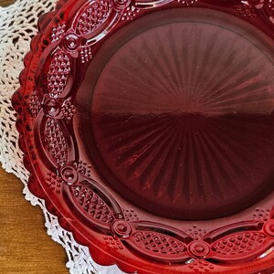 May include: A round, deep red glass plate featuring a scalloped edge and a decorative border with a pressed diamond pattern. The center of the plate displays a radiating sunburst design, and the overall surface is smooth and translucent.