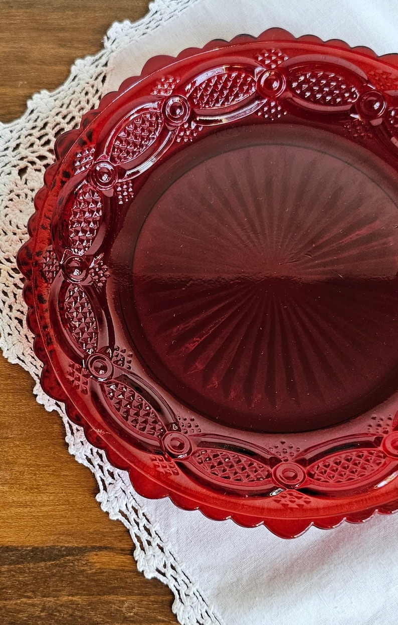 May include: A round, deep red glass plate featuring a scalloped edge and a decorative border with a pressed diamond pattern. The center of the plate displays a radiating sunburst design, and the overall surface is smooth and translucent.