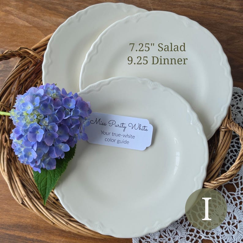 May include: Two off-white ceramic plates with scalloped edges, featuring text indicating sizes of 7.25 inches for salad and 9.25 inches for dinner. A blue hydrangea flower rests on the plates alongside a tag labeled Miss Purity White.