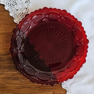 May include: A round, deep red glass plate featuring a scalloped edge and a decorative border with embossed geometric patterns. The center of the plate displays a sunburst design with radiating lines extending outward from the middle point.