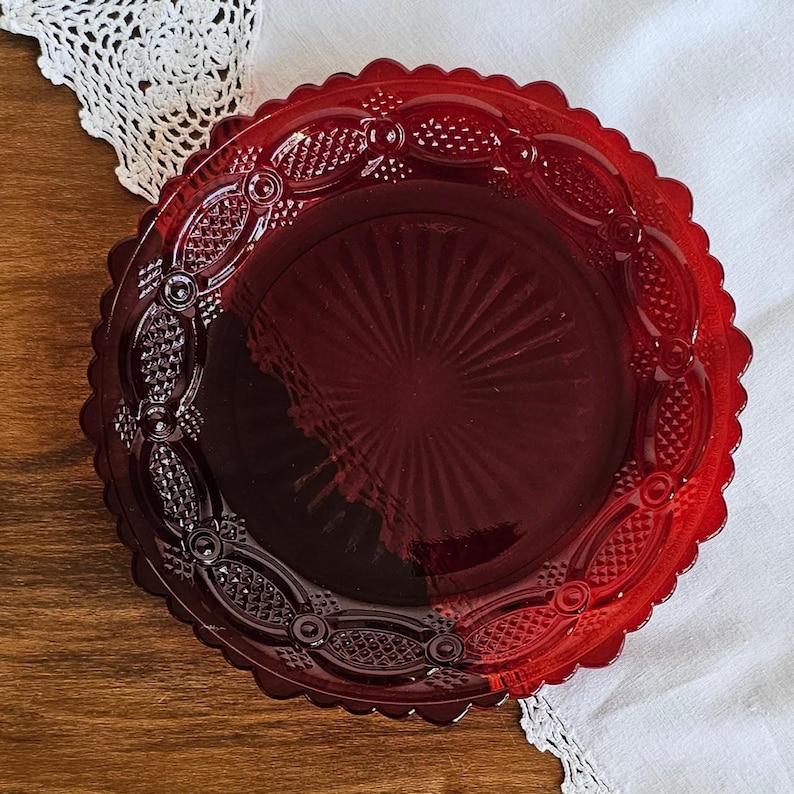 May include: A round, deep red glass plate featuring a scalloped edge and a decorative border with embossed geometric patterns. The center of the plate displays a sunburst design with radiating lines extending outward from the middle point.