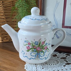 May include: White ceramic teapot with a lid, featuring a floral bouquet design with pink and blue tulips, green leaves, and blue ribbons. The teapot has a curved handle and spout, and a small knob on the lid. A decorative piece for serving tea.