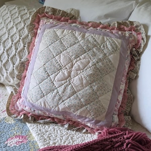 May include: Square quilted pillow with a floral pattern in soft pastel colors. The pillow has a light pink border and a ruffled edge in shades of pink and beige. The pillow is on a bed with other pillows and a blanket.