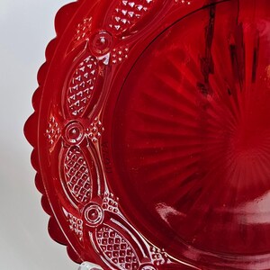 May include: A vibrant red glass plate featuring a scalloped edge and an intricate embossed pattern along the rim. The center of the plate displays a sunburst design with radiating lines, while the border includes textured diamond shapes.