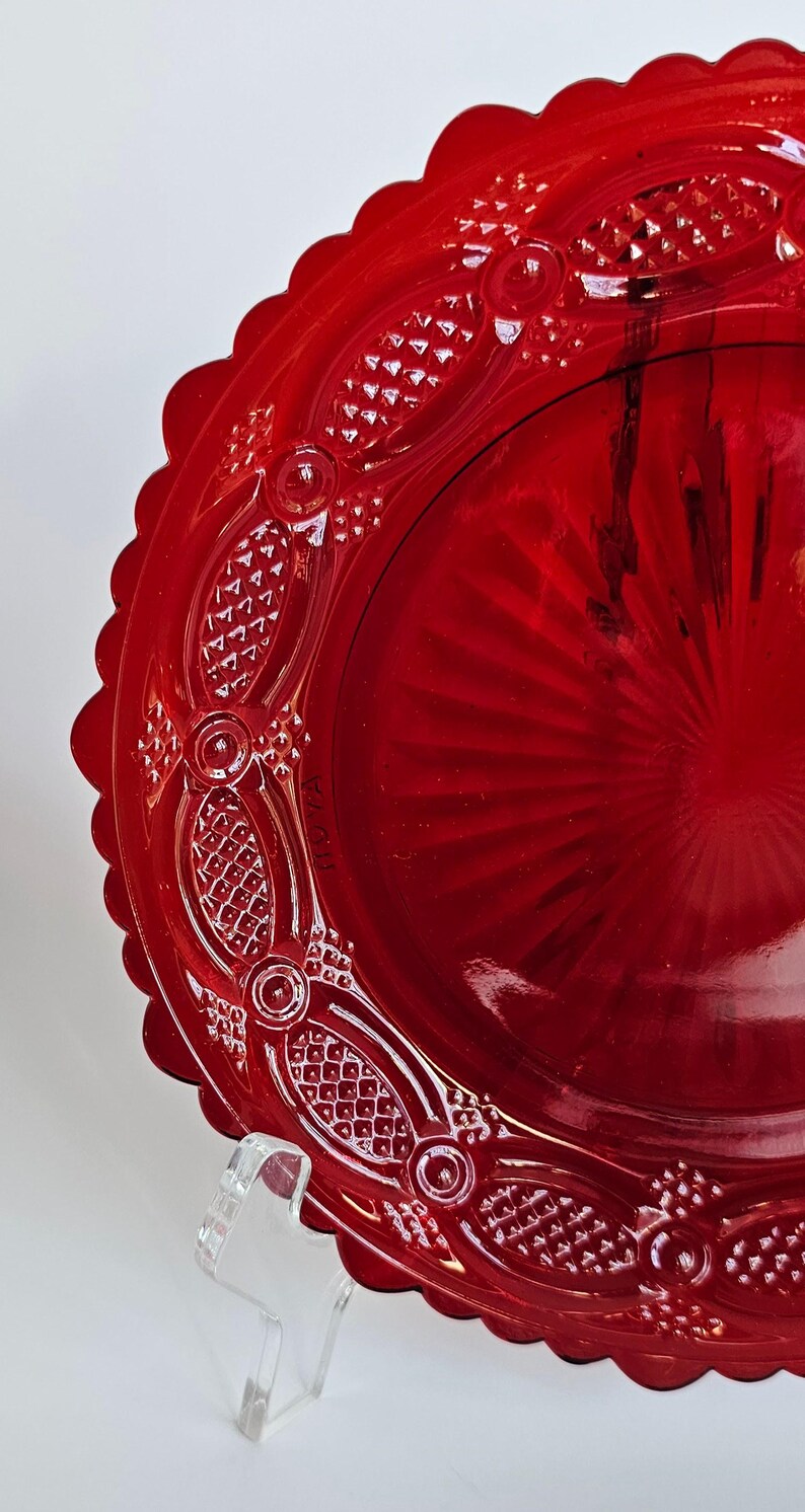 May include: A vibrant red glass plate featuring a scalloped edge and an intricate embossed pattern along the rim. The center of the plate displays a sunburst design with radiating lines, while the border includes textured diamond shapes.