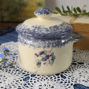 May include: A cream-colored ceramic jar with a matching lid, featuring a blue sponge-painted rim and a central botanical illustration of blackberries, leaves, and small white flowers. The jar has a single side handle and a rounded lid knob.
