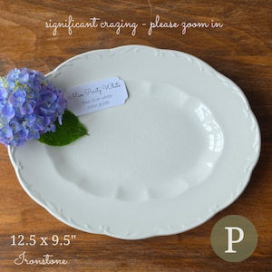 May include: An oval white ironstone platter with a scalloped edge and visible surface crazing. A small tag reads Miss Purity White Your true-white color guide. The platter measures 12.5 inches by 9.5 inches and features a blue hydrangea flower.