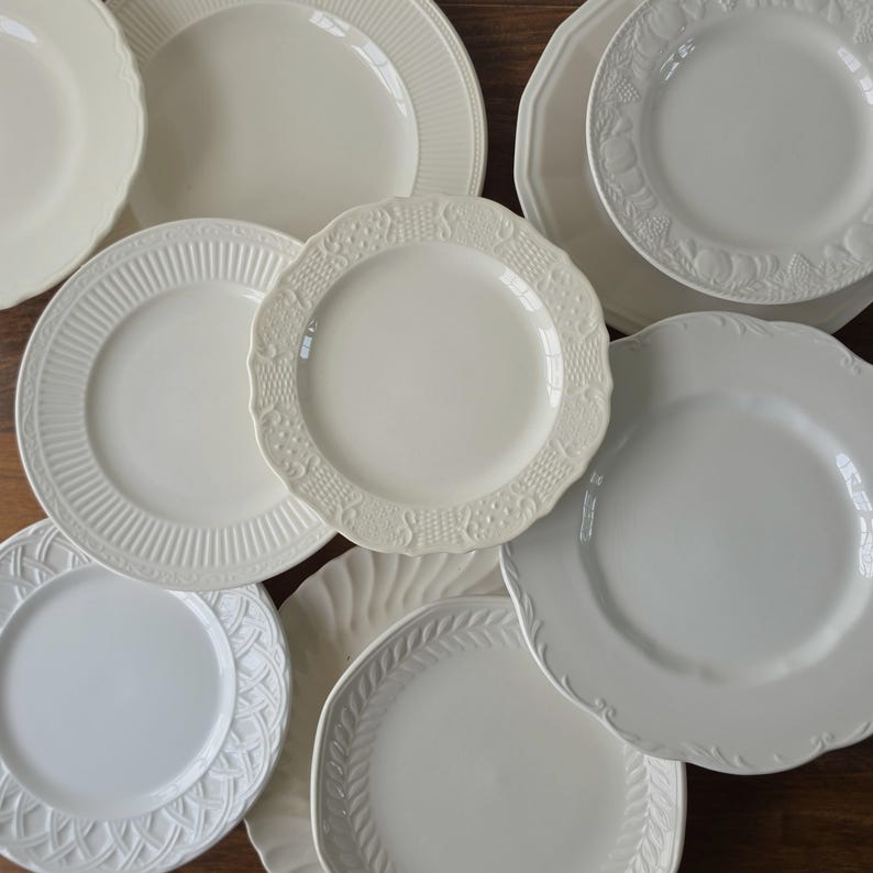 May include: A collection of cream and off-white ceramic plates featuring various embossed rim patterns, including basketweave, scalloped edges, fruit motifs, and vertical ribbing, arranged in an overlapping display of round dinnerware.