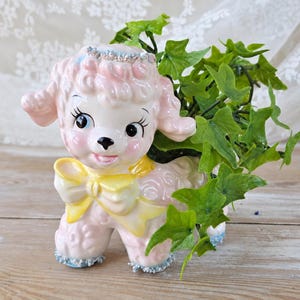 May include: A ceramic poodle figurine planter with a pale pink coat, a bright yellow bow around its neck, and blue glitter accents on its head and paws. The poodle has large dark eyes and is filled with green artificial ivy leaves.