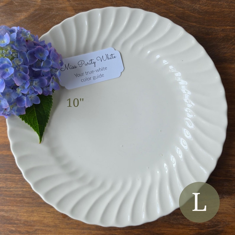 May include: A 10-inch white ceramic plate with a scalloped, wavy edge design. A small white tag is attached to the plate with the text Miss Purity White Your true-white color guide. A cluster of blue hydrangea flowers rests on the left side.