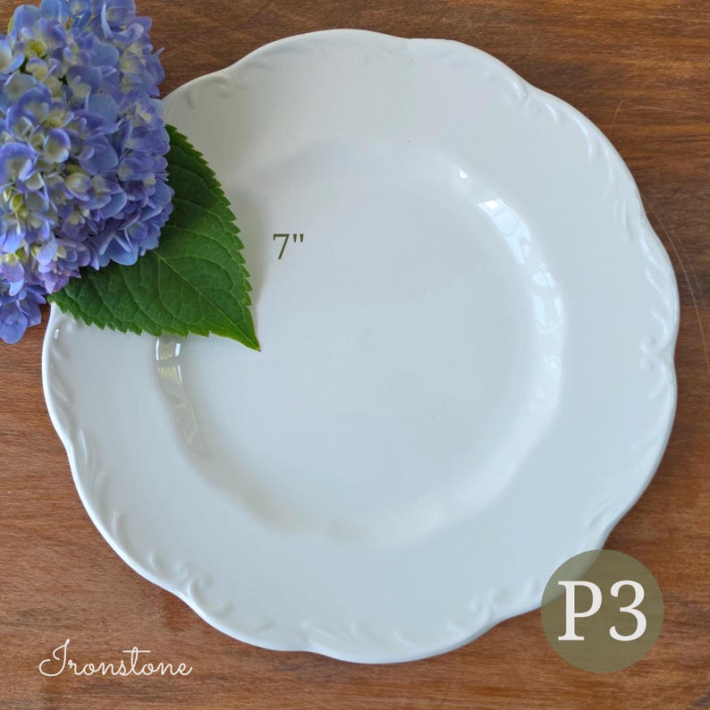 May include: A white ironstone plate with a scalloped edge and a 7-inch diameter, accompanied by a cluster of blue hydrangea flowers with green leaves. The plate features the text 7" and P3 printed on its surface, with the word Ironstone below.