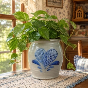 May include: A light gray ceramic crock featuring a blue hand-painted heart design with cross-hatch detailing. The vessel contains a lush, trailing pothos plant with vibrant green and variegated heart-shaped leaves spilling over the rounded rim.