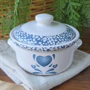 May include: A white ceramic casserole dish with a lid, featuring a blue heart design and speckled blue trim. The dish has two handles and a knob on the lid. The casserole dish is sitting on a white cloth.