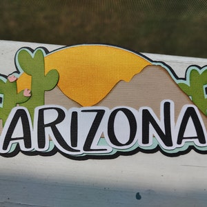 May include: A paper craft with a sunset, mountains, and cacti. The word "ARIZONA" is written in black letters on a white background.