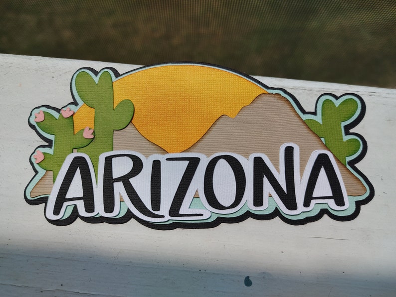 May include: A paper craft with a sunset, mountains, and cacti. The word "ARIZONA" is written in black letters on a white background.