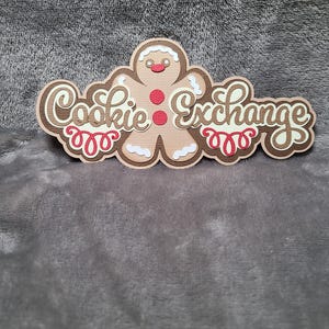 May include: A layered paper cutout featuring a gingerbread man design with the words "Cookie Exchange". The gingerbread man has a red mouth and buttons, with white icing details. The text is in a decorative font with brown and red accents.