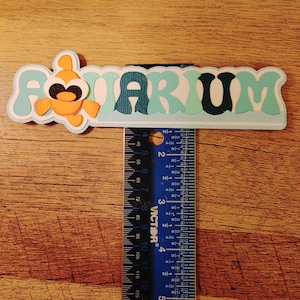 May include: A blue and white paper craft with the word "AQUARIUM" in a wavy font. An orange fish with black eyes is on the left side of the craft. A ruler is used to show the size of the craft, which is approximately 6 inches long.