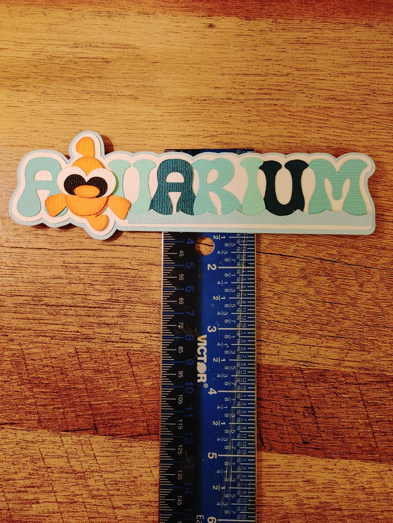 May include: A blue and white paper craft with the word "AQUARIUM" in a wavy font. An orange fish with black eyes is on the left side of the craft. A ruler is used to show the size of the craft, which is approximately 6 inches long.
