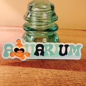 May include: A teal blue and white paper cutout of the word "Aquarium" with a cartoon orange fish with black eyes. The cutout is in front of a clear glass insulator.