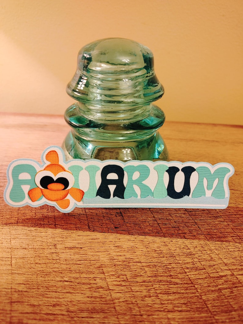 May include: A teal blue and white paper cutout of the word "Aquarium" with a cartoon orange fish with black eyes. The cutout is in front of a clear glass insulator.