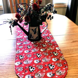 May include: Red and white table runner with a pattern of dogs wearing red, white, and blue hats and holding American flags. The runner is on a wooden table with a black watering can filled with red, white, and blue flowers.