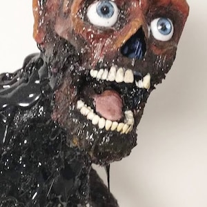 May include: A grotesque sculpture of a decaying figure with a skull-like face. The head is brown with blue eyes and an open mouth revealing teeth and a pink tongue. The body is black and appears to be covered in a shiny, viscous substance.