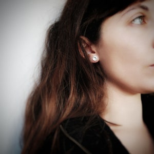 Puede incluir: Primer plano de la oreja de una mujer con un pequeño pendiente redondo de plata. El pendiente tiene una superficie texturizada y un centro ligeramente elevado.