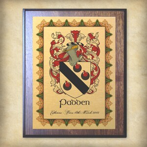 K&ouml;nnte beinhalten: Eine goldene Plakette mit einem braunen Holzrahmen. Die Plakette zeigt ein Wappen mit einem schwarz-wei&szlig;en Design, umgeben von roten und gr&uuml;nen floralen Akzenten. Der Text "Padden" ist unter dem Wappen gedruckt, gefolgt von "Catherine - Geboren am 18. M&auml;rz 2012".