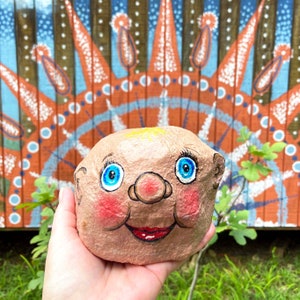 May include: A hand-painted rock with a smiling face. The rock is painted to look like a person with blue eyes, a red nose, and a red mouth. The rock is being held in a hand.