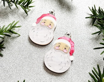May include: Two Santa Claus ornaments with pink hats and white beards. The ornaments have a silver clip at the top and are set against a silver background with green foliage. These festive ornaments are perfect for Christmas.