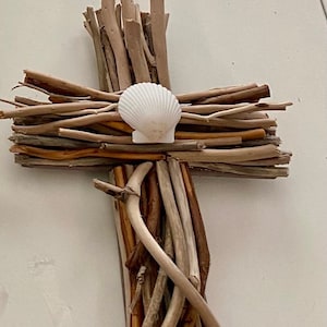 May include: A wooden cross made of driftwood branches with a white seashell in the center.