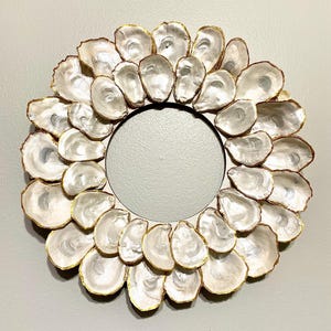 May include: A white oyster shell wreath with gold accents. The wreath is made of individual oyster shells that are arranged in a circular pattern. The shells are all white with a slight iridescent sheen. The gold accents are on the edges of the shells, giving the wreath a touch of elegance.