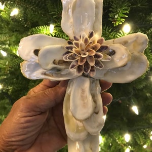 May include: A white oyster shell cross with a purple and brown seashell flower in the center. The cross is held in a hand against a green background.