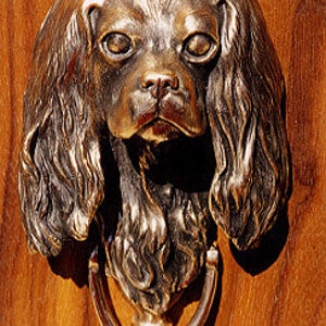May include: A bronze door knocker shaped like a Cavalier King Charles Spaniel dog's head. The knocker has a realistic design with a detailed face and flowing fur.