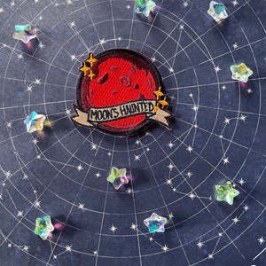 May include: A red embroidered patch with a crescent moon and the text "Moons Haunted" in a banner. The patch is surrounded by iridescent stars on a dark blue background.