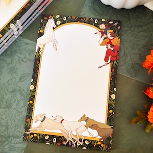 May include: A rectangular notepad featuring a white central area framed by a dark border with floral patterns. The design includes a white horse, three figures holding spears, and four running greyhound dogs in a meadow setting.