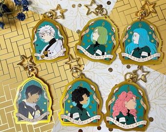 May include: Six gold-colored keychains featuring anime-style character portraits. Each keychain has a teal background with a gold border and a star-shaped charm. The characters have different hair colors and outfits. The text "Witch Hat Atelier" is visible.