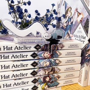 May include: A stack of "Witch Hat Atelier" manga volumes with a clear acrylic figure of a white creature and a spilled ink bottle. The volumes are numbered 1-6, with colorful illustrations.